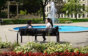 两个学生坐在费舍尔喷泉附近的长凳上, 用鲜花, trees, 喷泉, 建筑物照在阳光下, 夏天的一天.